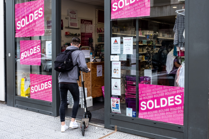 Un passant entre dans un magasin avec une trotinette. Sur la vitrine sont apposées cinq affiches roses indiquant : "soldes"