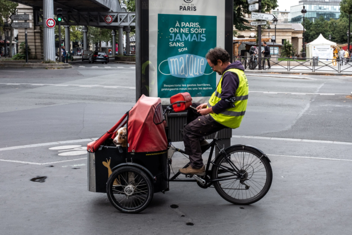 Un homme sur un tricycle, avec son chien, sans masque, devant un panneau indiquant qu'à Paris on ne sort jamais sans masque.